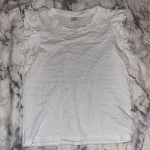 Vit volangtopp från Lindex - Säljer en vit topp från Lindex med volangdetaljer på axlarna. Toppen har en lätt strukturerad yta och är ärmlös med rund halsringning. Perfekt för dig som gillar en söt och enkel stil.