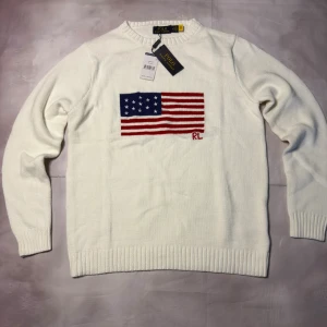 Vit stickad tröja Polo Ralph Lauren - Vit stickad tröja från Polo Ralph Lauren med amerikansk flagga broderad på bröstet och RL-detalj. Tröjan har rund hals, ribbade muddar och är långärmad. Materialet är mjuk bomull som känns skönt mot huden. Perfekt för dig som gillar klassisk och clean stil.