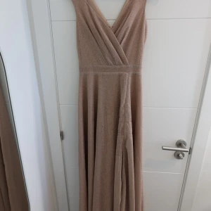 Glittrig rosafärgad långklänning - Elegant långklänning i rosafärgat glittrigt tyg med omlottliv och v-ringning. Klänningen är ärmlös och har en diskret slits framtill samt dragkedja i ryggen. Perfekt för dig som vill sticka ut med en lyxig och stilren look. Aldrig använd