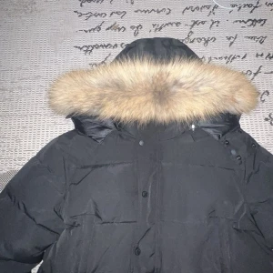 Svart dunjacka med pälsluva - Säljer en svart dunjacka med stor fluffig päls på luvan. Jackan har knappar framtill och är perfekt för kalla dagar. Den har en klassisk pufferstil och luvan ger extra värme och stilpoäng.