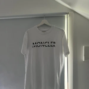 Vit Moncler t-shirt med logga - Säljer en vit t-shirt från Moncler med svart, glansig logotyp tryckt över bröstet och en svart patch på ärmen. Klassisk rund hals och korta ärmar. Materialet är mjuk bomull som känns skönt mot huden. Perfekt för dig som gillar stilrena designer och exklusiva märken.
