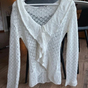 Vit stickad kofta från Saint Tropez - Supersöt vit stickad kofta från Saint Tropez i storlek M. Koftan har ett spetsigt mönster, volang längs kanten och en liten knytning framtill. Långärmad och lätt transparent, perfekt för lager-på-lager. Snygg detalj med vågiga kanter vid ärmslut och nederkant.