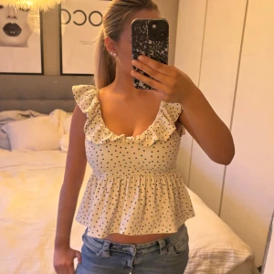 Prickig volangblus - Supersöt blus i ljus beige med svarta prickar och volangdetaljer vid axlarna. Blusen har en v-ringad front, ärmlös design och en öppen rygg med knytband. Perfekt till jeans eller kjol för en trendig look.