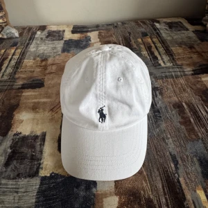 Vit Polo Ralph Lauren keps - Vit keps från Polo Ralph Lauren med klassisk böjd skärm och justerbart spänne bak. Broderad Polo-logga framtill och texten 'POLO' baktill. Tillverkad i bomull, perfekt för en clean och sportig look.