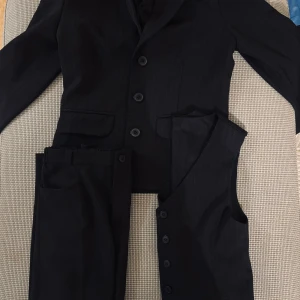 Barn Svart kostym med väst och byxa - Stilren svart kostym med kavaj, matchande väst och byxa. Kavajen har klassisk krage, tre knappar och fickor framtill. Västen har fem knappar och är ärmlös. Kostymen har en diskret randig struktur och passar perfekt till formella tillfällen.