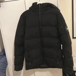 Supply and demand vinter jacka.  - Svart pufferjacka från Supply med stor huva och tydlig logga på ärmen. Jackan har dragkedja framtill, två sidofickor med dragkedja och SUPPLY-text på huvan. Perfekt för kalla dagar och har en clean, modern look. Pris kan diskuteras 