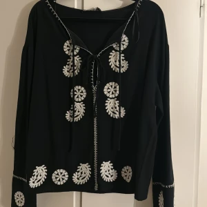 Svart blus med broderi från Zara - Svart blus från Zara med vita broderade detaljer och snörning framtill. Blusen har lång ärm och dekorativa sömmar längs halsringningen och ärmsluten. 
