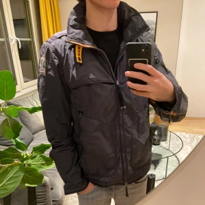 Grå vindjacka från Parajumpers - Snygg grå vindjacka från Parajumpers med hög krage, gul detalj vid halsen och patch på vänster ärm. Jackan har flera fickor, dragkedja framtill och är tillverkad i lätt polyester. Perfekt för dig som gillar stilren och sportig look. Storlek large men sitter som en medium 