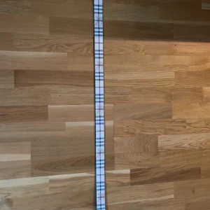 Rutig Burberry bälte med guldspänne - Snyggt bälte från Burberry med klassiskt rutigt mönster i vitt, brunt och svart. Bältet har ett stort guldigt spänne med Burberrys ikoniska TB-logo. Tillverkat i skinn med exklusiv känsla och stilren design. Storleken är 120cm