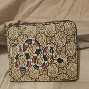 plånbok med ormmotiv - Snygg plånbok med klassiskt GG-mönster i beige och brunt, prydd med en cool orm i svart, rött och vitt på framsidan. Insidan har flera fack för kort och mynt, och är gjord i svart skinn. Perfekt för dig som gillar lyxiga detaljer och unik design.