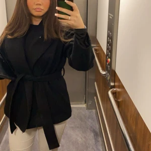 Svart omlott kappa med bälte - Säljer en svart kappa med omlott-design och bälte i midjan. Jackan har en loose passform och långa ärmar, perfekt för dig som gillar stilrena och enkla plagg. Materialet känns mjukt och har en klassisk look som funkar till jeans eller kostymbyxor.köpt för 800kr säljer för halva priset även fast den är i nästan helt nytt skick. 