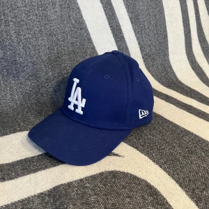 Blå LA  keps från New Era - Knappt använd. Snygg blå keps från New Era med LA Dodgers-logga broderad i vitt framtill. Klassisk 9SEVENTY-modell med böjd skärm och justerbar snapback baktill. Kepsen är tillverkad i bomull och har ventilerande hål för extra komfort.