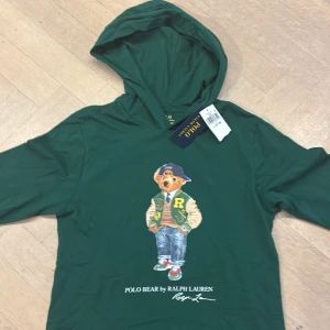 Grön hoodie Polo Bear Ralph Lauren - Snygg grön hoodie från Ralph Lauren med Polo Bear-tryck på bröstet. Tröjan har huva och långa ärmar, tillverkad i mjuk bomull. Perfekt för dig som gillar streetstyle och ikoniska motiv. Märkeslapp kvar och coolt print framtill. Helt ny, aldrig använd. Den är ganska liten i storlek. Pris kan diskuteras.