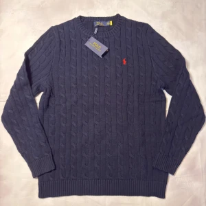 Mörkblå kabelstickad tröja Polo Ralph Lauren - Kabelstickad mörkblå tröja från Polo Ralph Lauren med klassisk rund hals och röd broderad logga på bröstet. Tillverkad i mjuk bomull och har ribbade muddar vid ärmslut och nederkant. Perfekt för dig som gillar stilren och tidlös design.