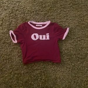 Vinröd croppad topp från Stay - Säljer en vinröd croppad topp från Stay med vit text 'Oui' framtill. T-shirten har korta ärmar och vita kontrastkanter vid hals och ärmslut. Passformen är tight. Perfekt för dig som gillar franska vibes och coola statement-plagg.