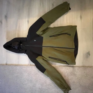 Grön och svart Skidjacka Cross Sportswear - Snygg vindjacka från Cross Sportswear i grön och svart färg med huva och dragkedja framtill. Jackan har två stora fickor med dragkedja och justerbar huva. Perfekt för dig som gillar outdoor och vill ha en cool look.