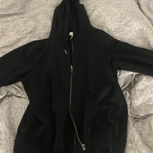 Svart hoodie med dragkedja - Svart hoodie med huva och snörning, hel dragkedja framtill och rymliga fickor. Perfekt för en chill och avslappnad stil. Klassisk modell med långa ärmar och mjukt material.