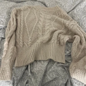 Beige stickad kabeltröja - Säljer en beige stickad tröja med grovt kabelstickat mönster och breda muddar. Tröjan har lång ärm och en mysig, oversized känsla. Perfekt för kyliga dagar när du vill vara både varm och snygg.