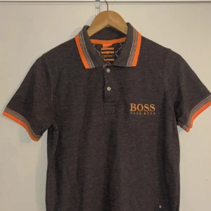  Hugo Boss piké  - Snygg grå pikétröja från Hugo Boss med orange logga på bröstet och coola orangea detaljer på krage och ärmslut. Storlek S men passar oxå M 