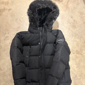 Supply & Demand Omega Longline Parka Junior - Säljer en svart pufferjacka från Supply & Demand med stor huva kantad av svart och grå fuskpäls. Jackan har dragkedja och knappar framtill, samt detaljerade band med text på framsidan och stora fickor. Perfekt för kalla dagar med sin tjocka vaddering, använd 2 gånger. I väldigt fint skick, köpt för 1000:- och priset går att diskutera vid snabb och smidig affär.