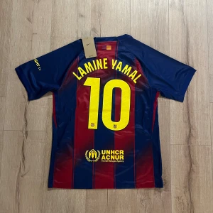 FC Barcelona Lamine Yamal tröja Nike S - Säljer en officiell FC Barcelona matchtröja från Nike med Lamine Yamal och nummer 10 på ryggen. Tröjan är kortärmad, har klassiska blå och röda vertikala ränder, gula detaljer och klubbmärke på bröstet. Materialet är lätt och ventilerande polyester.