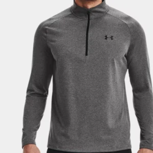 Grå långärmad träningströja från under armour - Snygg grå långärmad träningströja från Under Armour med svart logga på bröstet. Tröjan har en half zip-dragkedja och är gjord i mjukt syntetmaterial som andas. Perfekt för gymmet eller löprundan, stilren och sportig design.