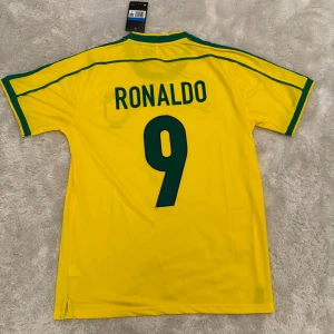 Brasilien Ronaldo 9 fotbollströja Nike - Gul Brasilien fotbollströja från Nike med gröna detaljer, nummer 9 och Ronaldo på ryggen. Klassisk design med landslagets emblem och Nike-logga på bröstet. Tillverkad i lätt och ventilerande material, perfekt för match eller träning.
