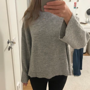 Grå stickad tröja med vida ärmar - Säljer en grå stickad tröja med breda, långa ärmar och ribbade muddar. Tröjan har en avslappnad passform och rund halsringning, perfekt för dig som gillar en chill och cozy stil. Enkel att matcha med jeans eller leggings.