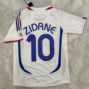 Frankrike Zidane 10 fotbollströja Adidas - Säljer en klassisk vit Frankrike fotbollströja från Adidas med Zidane och nummer 10 på ryggen. Tröjan har blå och röda detaljer, broderat landslagsmärke och Adidas-logga framtill. Tillverkad i lätt och ventilerande material, perfekt för fotbollsälskare.