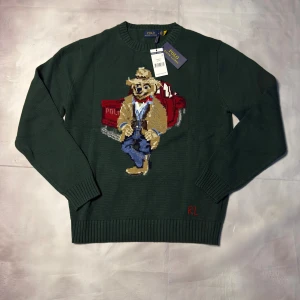 Grön stickad tröja Polo Ralph Lauren - Mörkgrön stickad tröja från Polo Ralph Lauren med broderad björn i cowboy-outfit och röd bil på framsidan. Rund hals, ribbade muddar och RL-broderi vid nederkanten. Tillverkad i bomull, perfekt för dig som gillar statement-plagg.