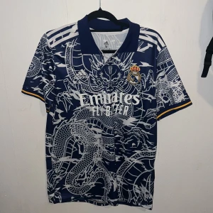 Real Madrid drakmönstrad fotbollströja - Snygg Real Madrid fotbollströja från Adidas i marinblått med vit drakmönster över hela tröjan. Broderat klubbmärke på bröstet och gula detaljer vid ärmslut. Klassisk krage och korta ärmar. Tillverkad i lätt och ventilerande polyester.