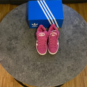 Rosa Adidas campus  - Säljer ett par snygga rosa Adidas sneakers med klassiska vita detaljer och snörning. Skorna har en rund tå och platt sula, perfekt för dig som gillar färgstarka och sportiga sneakers. Kommer med originalkartong. Storlek 39 1/3 HELT NYA! Aldrig använda. 