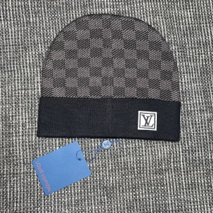 Louis Vuitton rutig mössa i svart - Snygg mössa från Louis Vuitton med klassiskt rutigt mönster i mörkgrått och svart. Mössan har en bred svart uppvikt kant med LV-logga framtill. Materialet känns mjukt och lyxigt, perfekt för att lyfta din stil under kyliga dagar.
