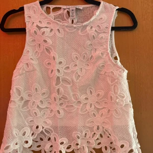 Vit blommig spetsblus från NLY Trend - Säljer en vit blus från NLY Trend i storlek 36. Blusen har ett blommigt spetsmönster med genombrutna detaljer och är ärmlös. Perfekt för dig som gillar romantisk och feminin stil. Materialet känns lätt och luftigt, och blusen har en rund hals.