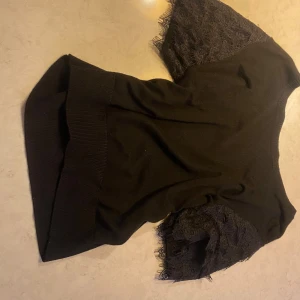 Svart topp med spetsärmar - Säljer en svart topp med bred ribbad kant nedtill och korta ärmar i snygg spets med fransiga detaljer. Toppen har en mjuk, stickad känsla i kroppen och ärmarna har ett blommigt spetsmönster som ger en cool kontrast. Perfekt för dig som gillar stilrena plagg med twist.