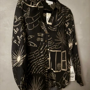 Skjorta - Svart skjorta med coolt handskrivet print och vita teckningar av palmer, fönster och text. Skjortan har klassisk krage, knappar framtill och lång ärm. Materialet är mjukt och flowy, perfekt för dig som gillar statement-plagg med unik vibe. Ord pris 1400kr