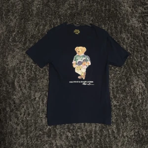 Mörkblå t-shirt med björntryck från Polo Ralph Lauren - Säljer en mörkblå t-shirt från Polo Ralph Lauren med ett stort tryck av en björn på framsidan. Klassisk rund halsringning och korta ärmar. Perfekt för dig som vill ha något unikt men stilrent i garderoben.
