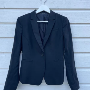 Tiger of Sweden kavaj - Klassisk svart kavaj från Tiger of Sweden. Klassisk design och enkel knappstängning. Perfekt för en stilren look. Kavajen har en smickrande passform och är tillverkad i ett bekvämt material. Strukturmönstrad med dold knapp vid ärmslut. 