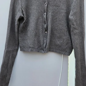 Grå ribbad kofta från Lager 157 storl.M - Grå croppad kofta från Lager 157 med ribbad struktur och långa ärmar. Tröjan har knappar framtill och är tillverkad i ett mjukt stickat material som känns skönt mot huden. Perfekt att slänga över en t-shirt för en chill look.