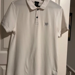 Vit slim fit t-shirt från Hugo Boss - Vit t-shirt från Hugo Boss med klassisk krage och två svarta knappar framtill. T-shirten har korta ärmar och en diskret Boss-logga på bröstet. Materialet är mjuk bomull som känns skönt mot huden och ger en clean look.