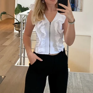 Vit blus med volanger och dragkedja - Säljer en fräsch vit blus med volanger längs fram och axlarna. Blusen har en snygg v-ringning och dragkedja framtill som ger en cool touch. Materialet känns lätt och passar perfekt till varmare dagar. Stilren och trendig topp för dig som gillar detaljer.