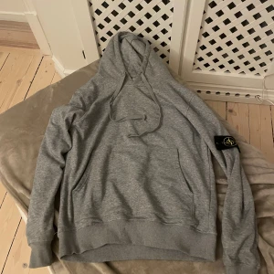 Grå hoodie från Stone Island - Snygg grå hoodie från Stone Island med klassisk logopatch på vänster ärm. Tröjan har huva med snörning, ribbade muddar och en stor magficka. Perfekt för chill dagar och streetwear-stil. Mjuk bomullskvalitet och normal passform.