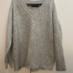 Grå stickad v-ringad tröja  - Priset kan diskuteras! Mysig grå stickad tröja med v-ringning och bred ribbad kant vid halsen. Tröjan har lång ärm och en relaxed passform, perfekt för lager på lager under kyliga dagar. Enkel och stilren design som funkar till allt.