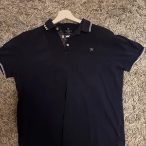 Mörkblå pikétröja från Hackett - Snygg mörkblå pikétröja från Hackett London med klassisk krage och knappar framtill. Tröjan har vita detaljer på krage och ärmslut samt diskret logga på bröstet. Perfekt för en stilren och avslappnad look.