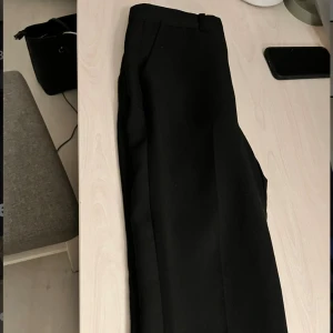 Svarta kostymbyxor med rak passform - Snygga svarta kostymbyxor med klassisk rak siluett och pressveck. Byxorna har midjehöjd med hällor för bälte och är tillverkade i ett slätt tyg som ger en stilren look. Perfekta för dig som gillar minimalistisk och clean stil.