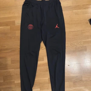 Svarta Jordan PSG träningsbyxor  - Svarta träningsbyxor från Jordan x PSG med röd logga och Paris Saint-Germain emblem på framsidan. Slim fit-modell med elastisk midja och smalare ben. Materialet är mjukt och stretchigt, perfekt för sport eller chill. Snygg och modern look med diskreta detaljer.