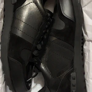 Valentino Garavani Rockrunners svarta - Svarta Valentino Garavani Rockrunner sneakers med coola camouflagedetaljer. Snygg mix av skinn och mocka, platt sula med nitar på sidan och rund tå. Klassisk snörning och diskret logga på plösen. Perfekta för dig som gillar stilrena och edgy skor. Helt nya ( Aldrig använda )