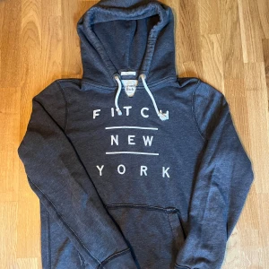 Abercrombie & Fitch hoodie  - Snygg mörkgrå hoodie från Abercrombie & Fitch med vit text 'FITCH NEW YORK' på bröstet. Tröjan har huva med snörning, ribbade muddar och en klassisk känguruficka framtill. Perfekt för chill dagar och streetstyle.