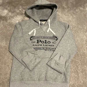 Grå hoodie från Polo Ralph Lauren - Snygg grå hoodie från Polo Ralph Lauren med svart tryck framtill. Tröjan har huva med vita snören, ribbade muddar och känguruficka. Tillverkad i 100% bomull för skön känsla. Perfekt för chill dagar nu mot vintern