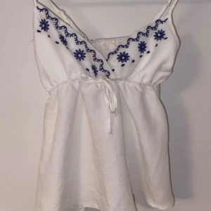Vit linnetopp med blå broderi - Aldrig använd. Supersöt vit linnetopp med tunna axelband och blått blommigt broderi längs v-ringningen. Toppen har knyt i midjan för en snygg passform och är tillverkad i ett lätt, luftigt material som passar perfekt till varma dagar. Broderidetaljerna ger en boho-vibe.